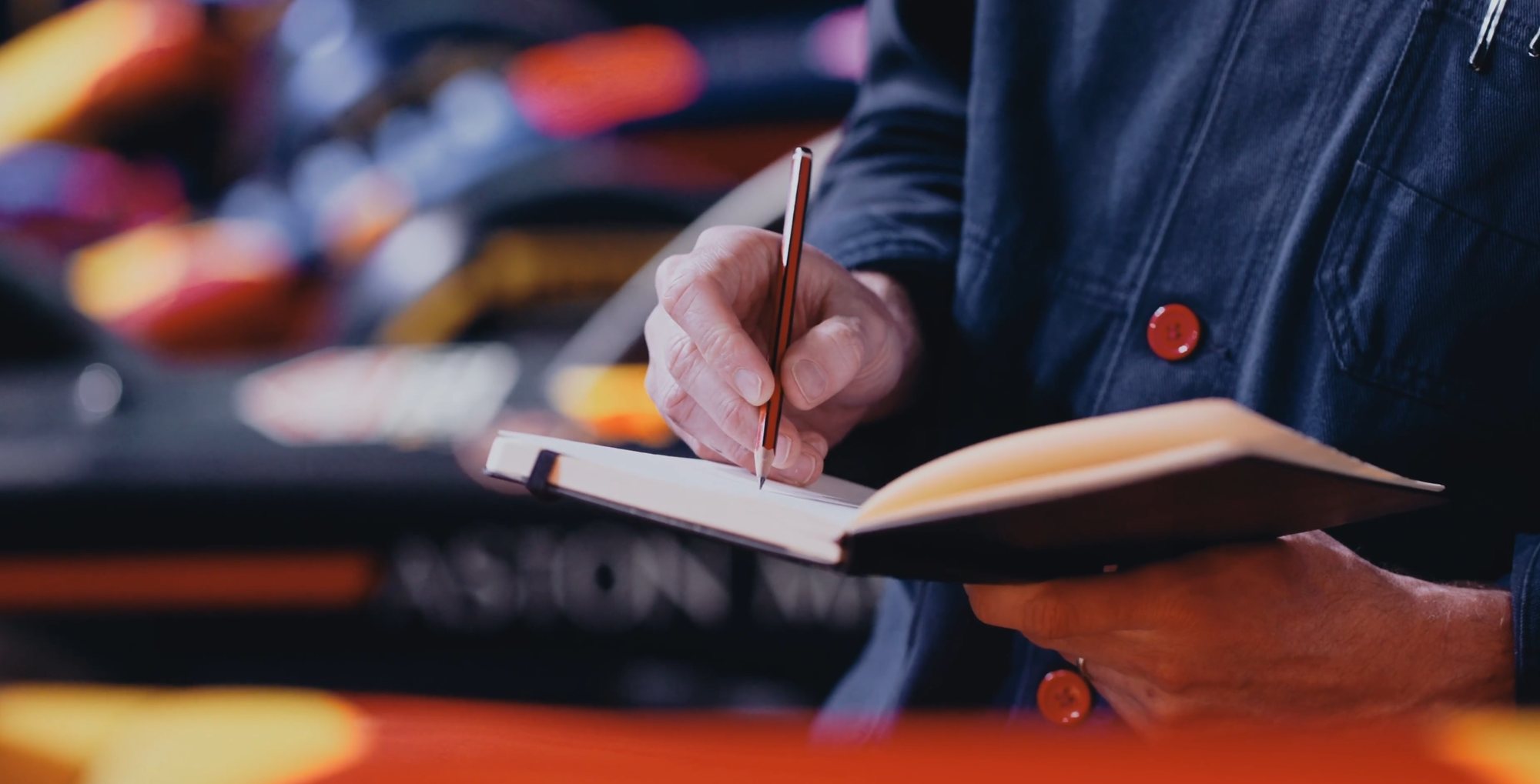 Close-up of a hand making notes with a pencil in a notebook, a race car and warm lights blurred behind.